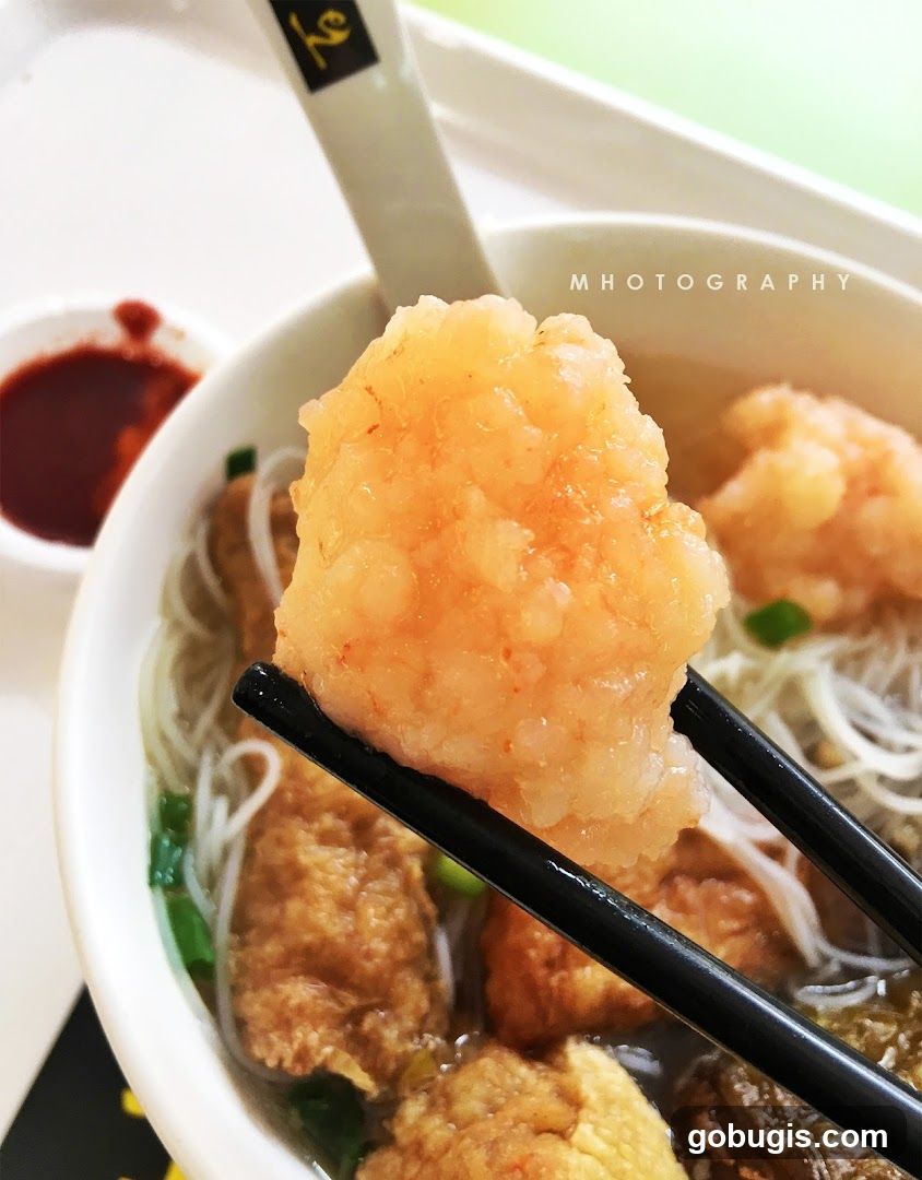 Close-up of a bowl of noodle soup with a large piece of shrimp being held by chopsticks.