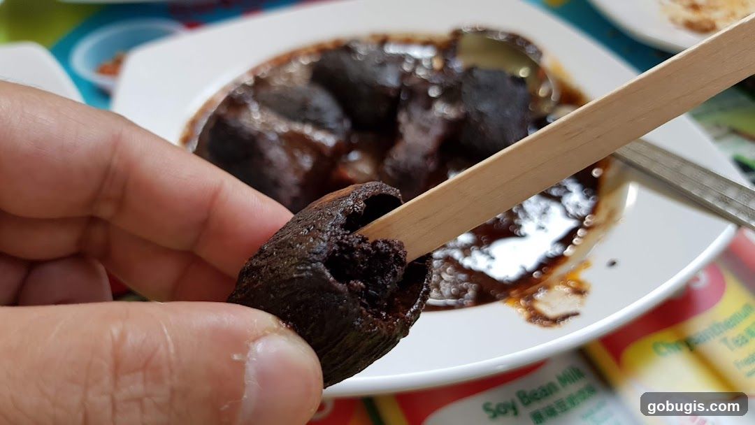 Close-up of a person holding a dark, hollowed-out food item on a wooden skewer, with a dish of similar items in a dark sauce in the background.