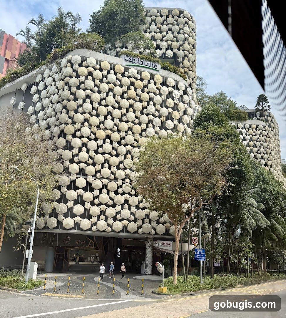 A view of the Capitaland building with its distinctive honeycomb-like facade. The building is adorned with numerous hexagonal concrete elements, creating a textured and visually striking exterior. Lush greenery cascades from the rooftops and is interspersed throughout the structure. The ground level features several entrances and storefronts, with people walking by. Signage for "24HRS KIN WANG CAFE" is visible on the right side of the entrance area.