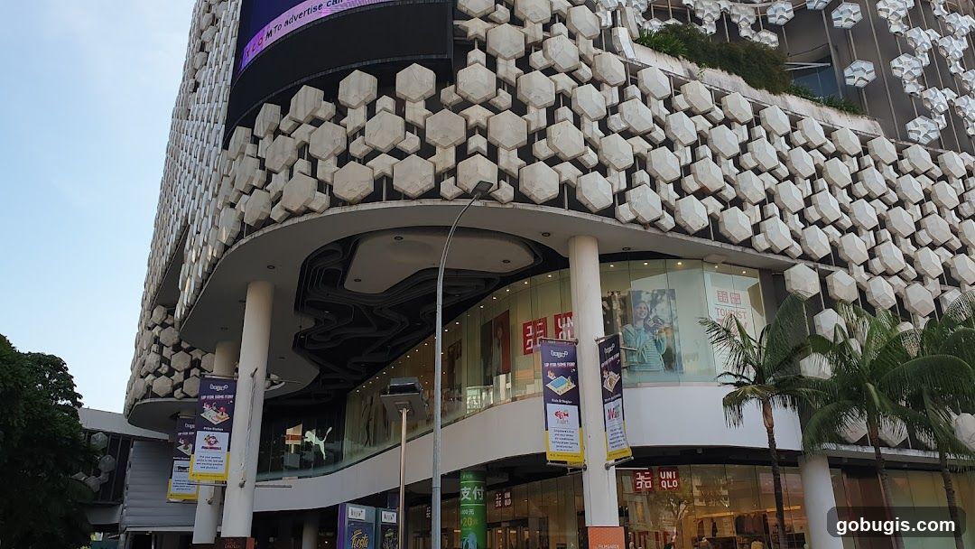 A wide shot of a modern building with a distinctive honeycomb-like facade. The lower levels feature large glass windows revealing store interiors. Prominent signage for UNIQLO is visible. Banners hang from the building, advertising events or promotions. Palm trees are in the foreground.