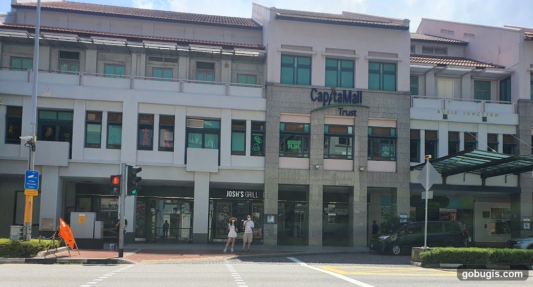 A street view of the exterior of a building complex with multiple shops. The building has a modern facade with large windows and signage. Visible signs include "CapitaMall Trust" and "BUGIS JUNCTION". Several storefronts are visible, including "JOSH'S GRILL" and "SUSHI PLUS". Two people are standing on the sidewalk in front of Josh's Grill.