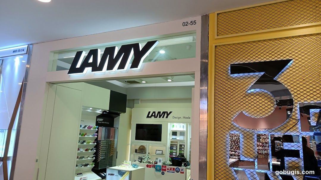 The exterior of a Lamy store with the Lamy logo prominently displayed above the entrance. Inside, shelves are stocked with pens and writing instruments. A large number '3' is visible on an adjacent wall.