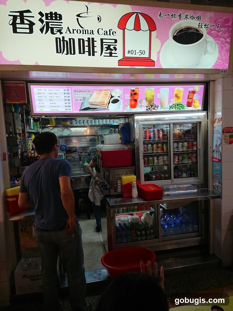 A street-level view of Aroma Cafe, a food stall with a prominent sign featuring Chinese characters and the English name "Aroma Cafe". Below the main sign is a menu board displaying various drinks and a breakfast set. The stall has a refrigerated display case filled with canned drinks and bottles. A customer is standing at the counter, and a staff member is visible behind it.