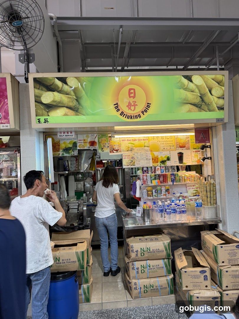A street-level view of a beverage stall with a prominent sign featuring sugarcane. A man is seen eating, and a woman is working behind the counter. Stacks of boxes are visible in front of the stall.