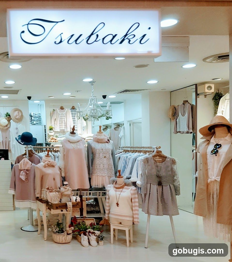 A well-lit clothing store interior with mannequins displaying women's apparel. The store name 'Tsubaki' is visible on a illuminated sign above the entrance. Racks of clothing are arranged throughout the space, with a decorative chandelier hanging from the ceiling.