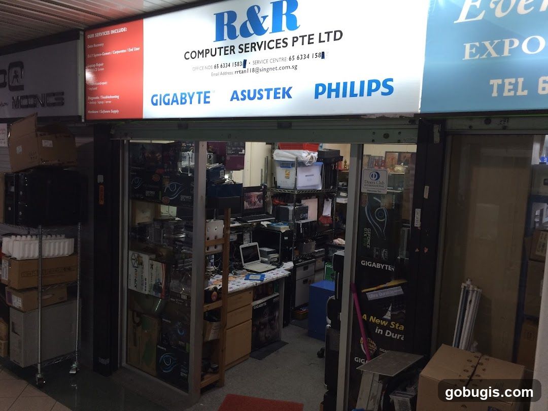 The storefront of R&R Computer Services Pte Ltd, a computer repair and supply shop. The sign above the entrance lists services such as Data Recovery, D.I.Y. System-Gamers/Corporates/End User, Laptop Repair, Troubleshooting, and Hardware/Software Supply. Brands like Gigabyte, Asus, and Philips are also advertised. The interior of the shop is visible, filled with computer parts and equipment.