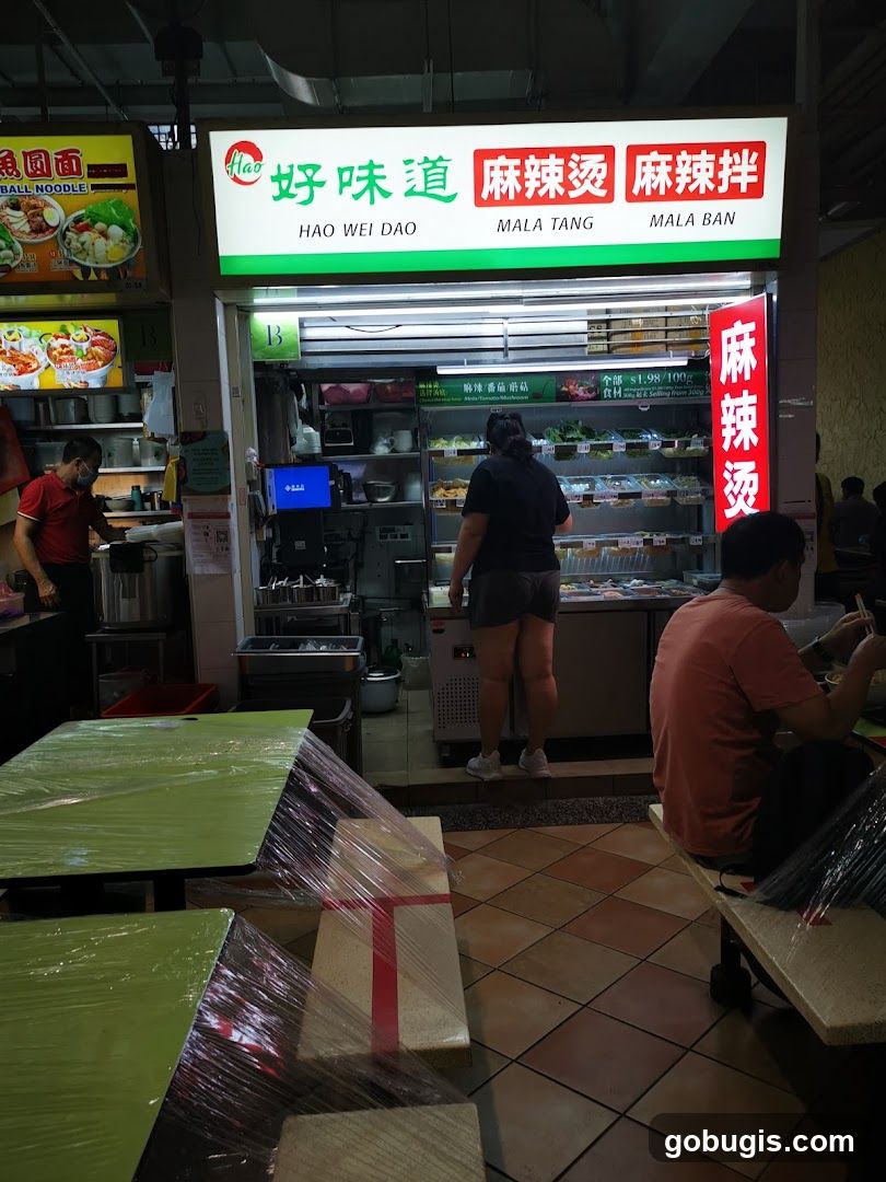 A food stall with a prominent sign displaying "Hao Wei Dao" in Chinese and English, along with "Mala Tang" and "Mala Ban". The stall offers a variety of ingredients displayed in refrigerated cases. A price of $1.98/100g is visible. Tables covered in plastic wrap are in the foreground.