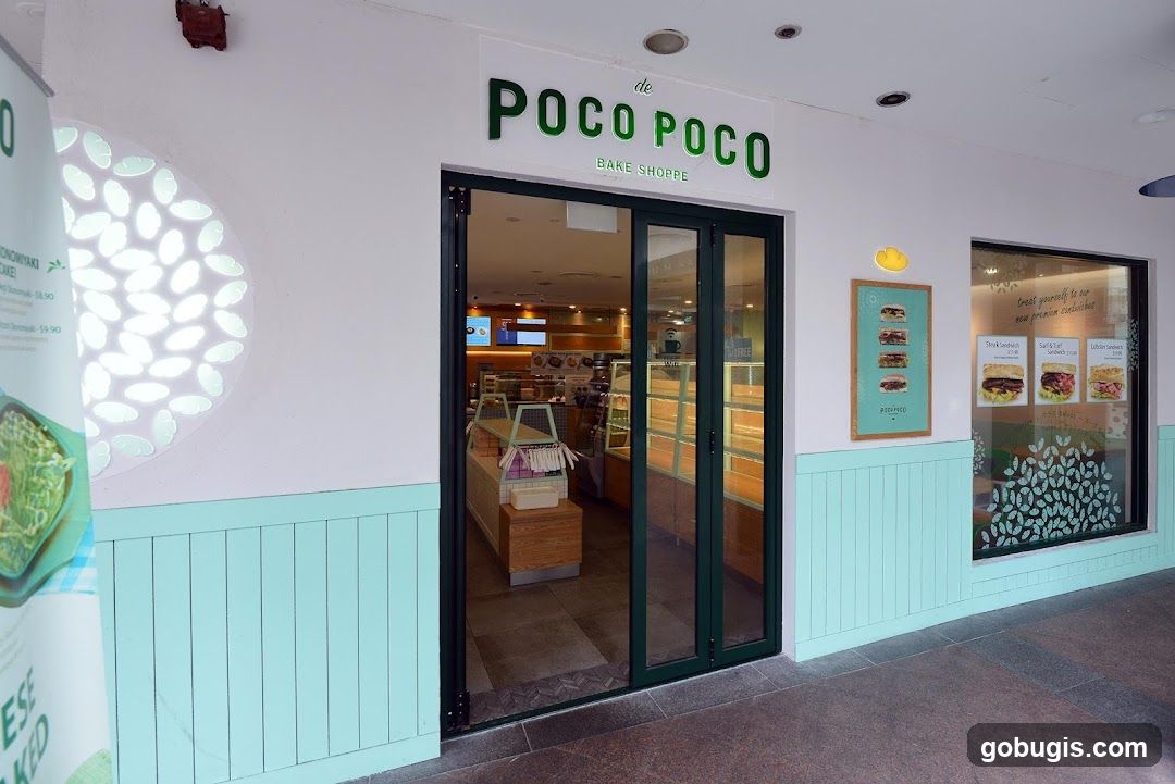The exterior of a bake shoppe with a green and white color scheme. The shop has large glass doors and windows. Above the entrance, the name "de POCO POCO BAKE SHOPPE" is displayed in green lettering. To the right of the entrance, a window displays images and prices of sandwiches. A menu board with images of sandwiches is also visible. A large circular light fixture with a cloud pattern is on the wall to the left of the entrance.