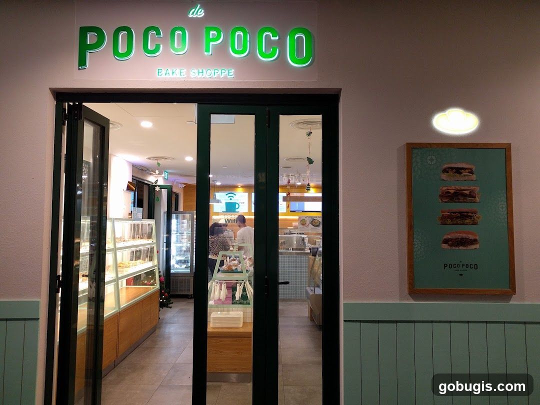 The exterior of a bake shoppe with a prominent green neon sign that reads "de POCO POCO BAKE SHOPPE". The entrance features double glass doors, with one set slightly ajar, revealing display cases filled with baked goods and a glimpse of the interior. To the right of the entrance, a framed poster displays four different sandwiches.