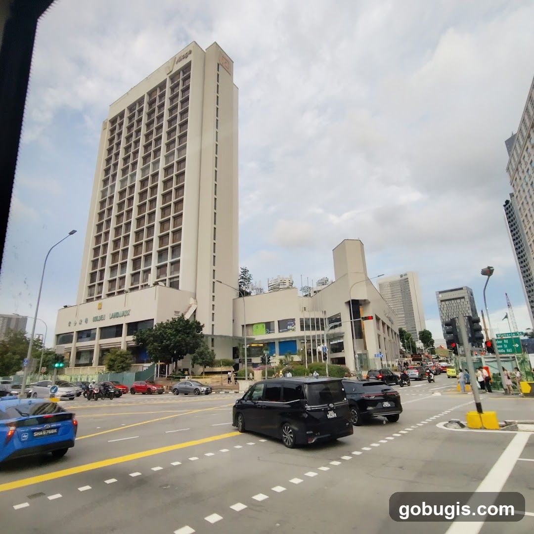 A tall, cream-colored hotel building with many windows stands prominently on a city street. The hotel's name, "GOLDEN LANDMARK," is visible in large letters on the lower facade. Below the hotel, there are several shops and businesses with glass storefronts. Cars and motorcycles are driving on the street in front of the building, and pedestrians are visible on the sidewalk. The sky is cloudy.