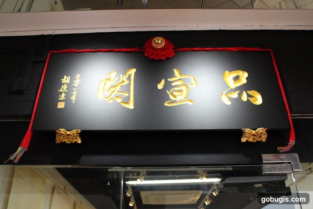 A black sign with gold Chinese characters and decorative elements hangs above a glass storefront. The sign features large, ornate gold lettering in the center, with smaller gold text and a red seal on the left. Red tassels and a red decorative ornament are attached to the top of the sign. Two small golden lion-like figures are mounted on the bottom corners of the sign.
