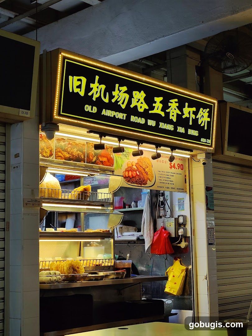 A brightly lit food stall with a prominent sign displaying Chinese characters and the English name "OLD AIRPORT ROAD WU XIANG XIA BING". The stall displays various fried food items behind glass. A menu board is visible with prices and item descriptions.