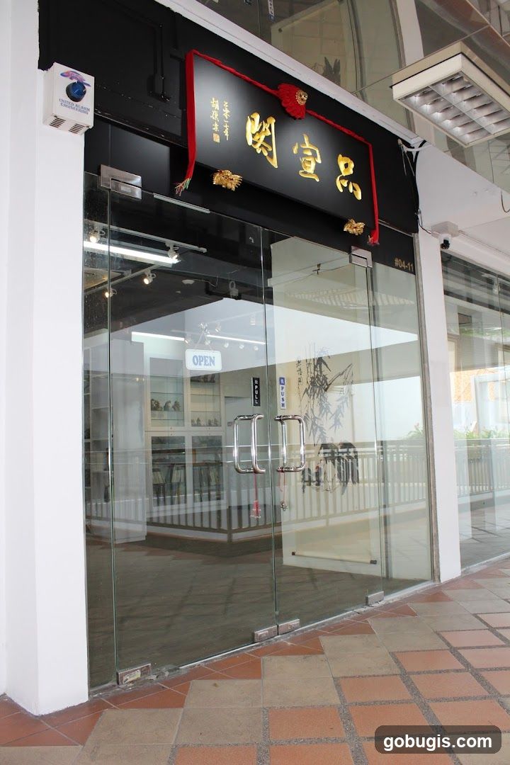 A modern storefront with large glass doors and windows. Above the entrance, a black sign with golden Chinese characters is displayed, adorned with red ribbons and decorative gold ornaments. A white sign indicating "OPEN" is visible through the glass doors. The interior shows display shelves with various items.