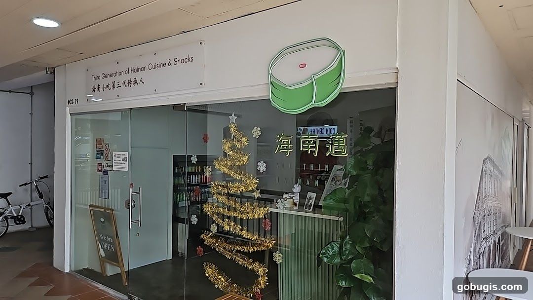The storefront of a restaurant with glass doors and windows. A sign above the entrance reads "Third Generation of Hainan Cuisine & Snacks" in English and Chinese. A decorative green and white graphic of a food item is displayed above the entrance. Inside, a Christmas tree decorated with gold tinsel and snowflakes is visible, along with plants and a counter. A bicycle is parked outside to the left, and an "OPEN" sign is displayed on an easel near the entrance.