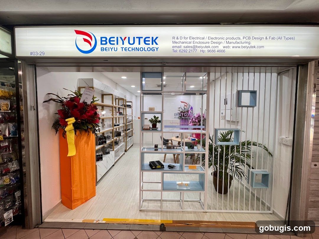 The storefront of Beiyutek, a company specializing in R&D for electrical/electronic products, PCB design and fabrication, and mechanical enclosure design/manufacturing. The shop front features a prominent illuminated sign with the company logo and name, along with contact information. Inside, shelves display various electronic components and products. A floral arrangement is placed near the entrance.