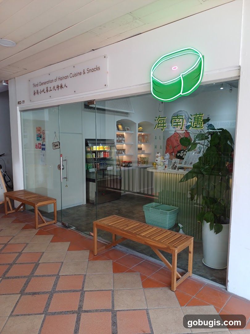 The exterior of a shop with a glass facade. A sign above the entrance reads "Third Generation of Hainan Cuisine & Snacks" in English and Chinese. A neon sign shaped like a green bun with a pink center is mounted above the door. Inside, shelves display food items, and a refrigerator is visible. Two wooden benches are placed outside the entrance.
