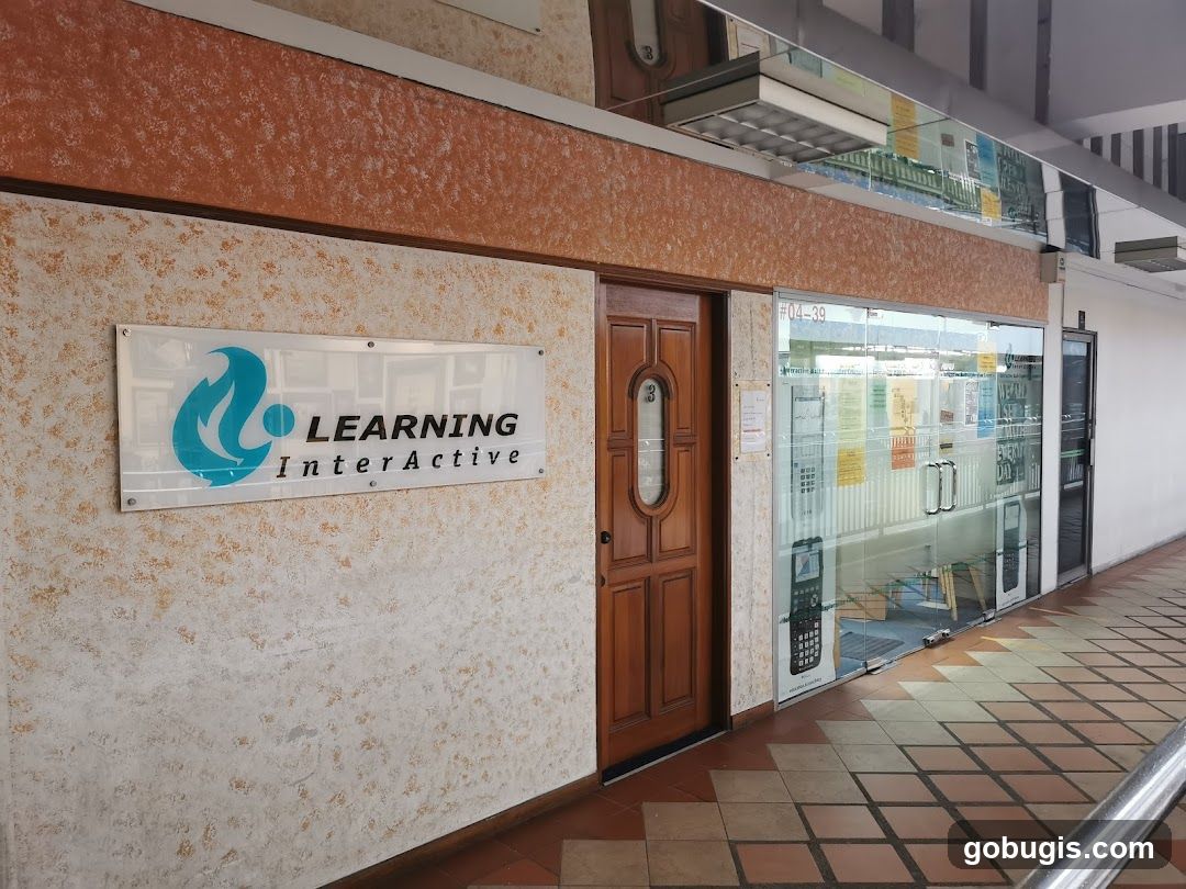 The exterior of a business with a sign that reads "LEARNING InterActive" and a logo. There is a wooden door to the left of the entrance and a glass storefront to the right. The storefront has various posters and signs displayed on and behind the glass. The floor is tiled in a diamond pattern.