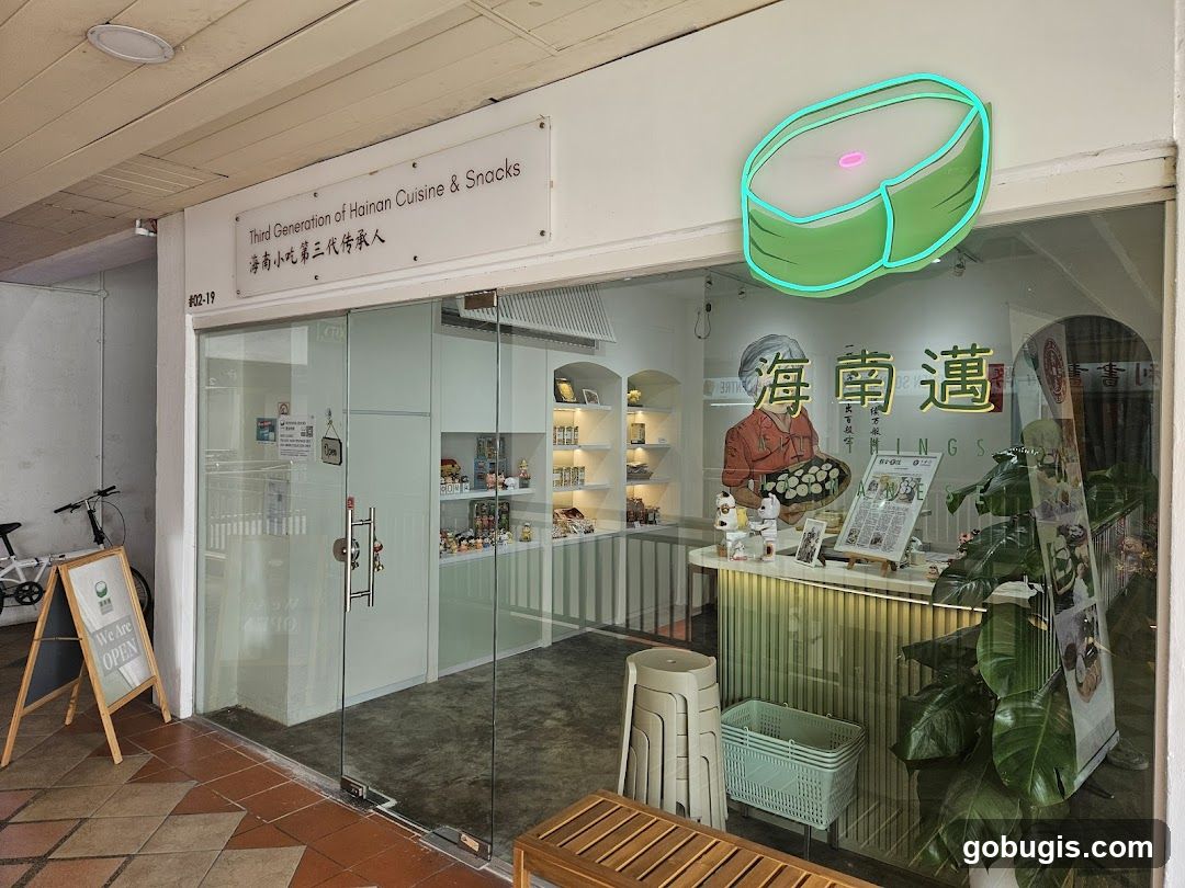 The exterior of a shop with large glass windows and doors. A sign above the entrance reads "Third Generation of Hainan Cuisine & Snacks" in English and Chinese. A neon sign shaped like a green dessert is visible above the entrance. Inside, shelves display various items, and a counter is visible. A bicycle is parked outside to the left, and a wooden A-frame sign with "We Are OPEN" is also present.