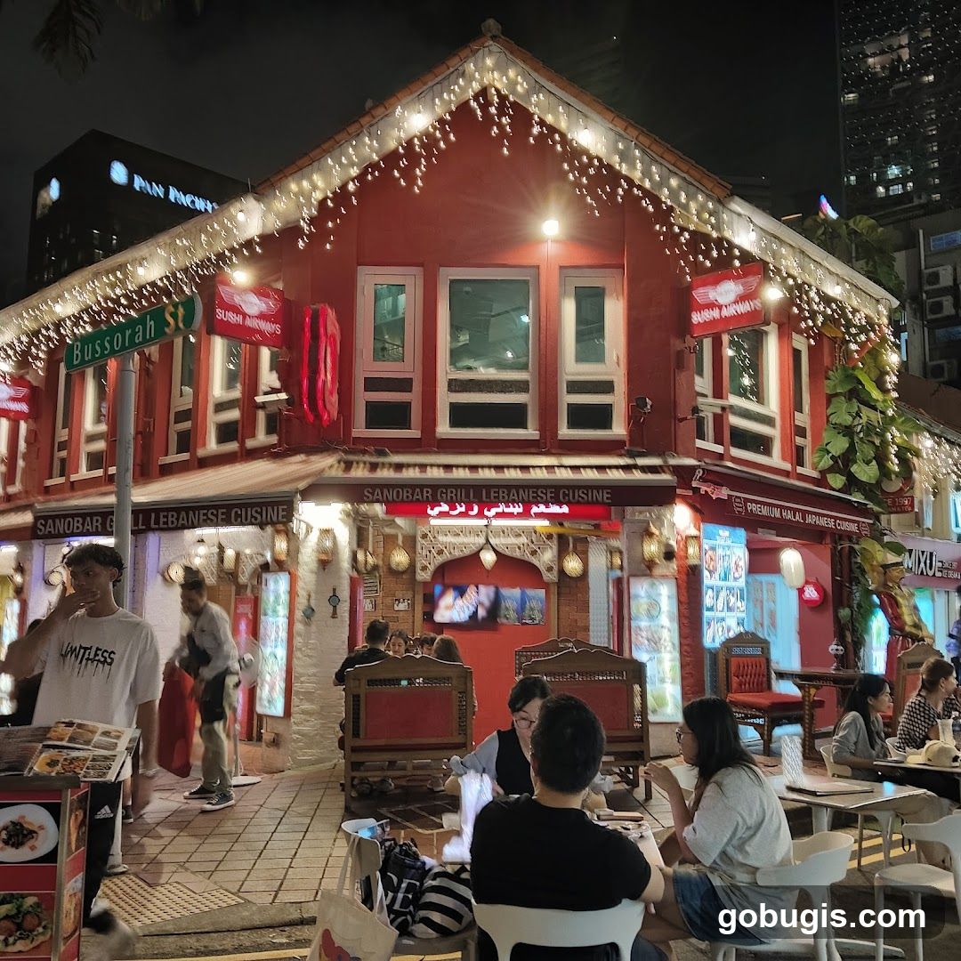 A red building with white trim and festive lights is a restaurant. Signs indicate it serves Lebanese and Japanese cuisine. People are seated at tables outside the restaurant.