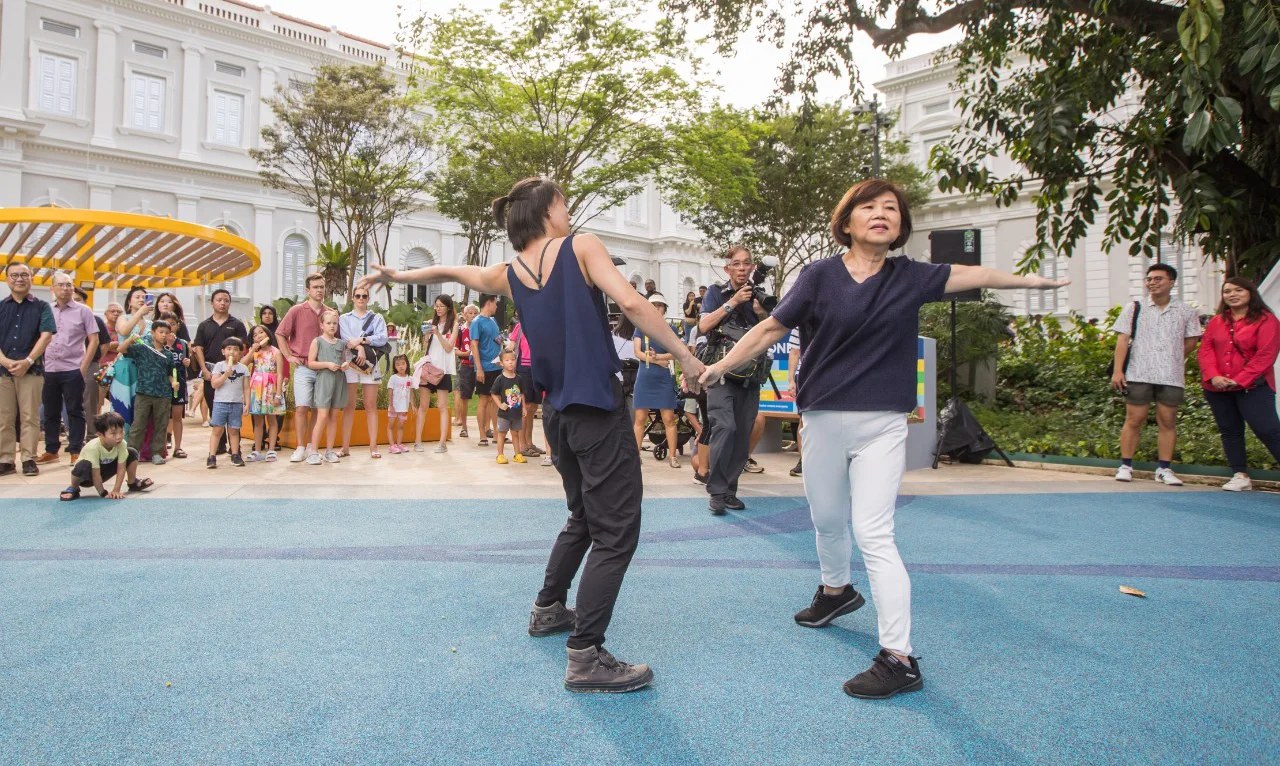Two women dancing on the blue playground surface of the National Museum front lawn, with the museum facade and a crowd of families behind them