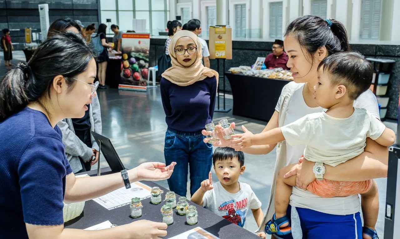 A child reaching for small glass jars at a National Museum marketplace booth, with a parent holding a toddler and a volunteer behind the table