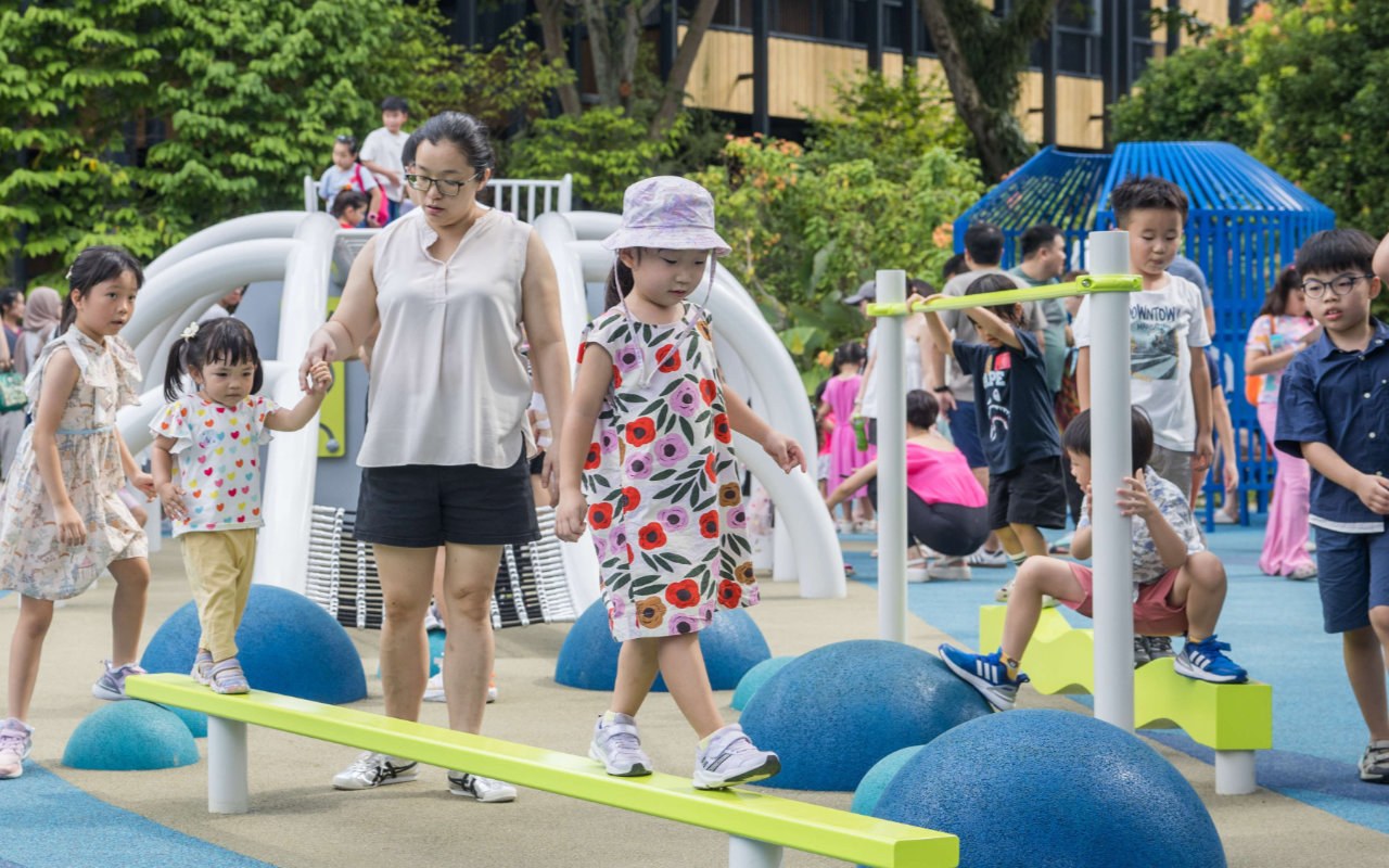 Children and an adult walking a lime-green balance beam on the National Museum front-lawn playground, with white climbing frames behind