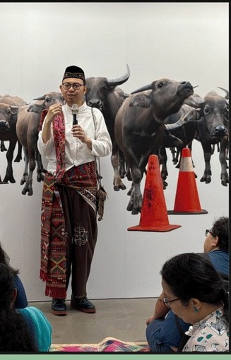 Storyteller Hafiz Rashid in a songkok and woven sash, microphone in hand, standing before a wall of water buffaloes at ArCH Square