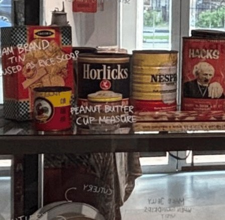 Vintage Horlicks, Nespray and Hacks tins on a glass shelf, reused as rice scoops and cup measures in the Museum of Food display